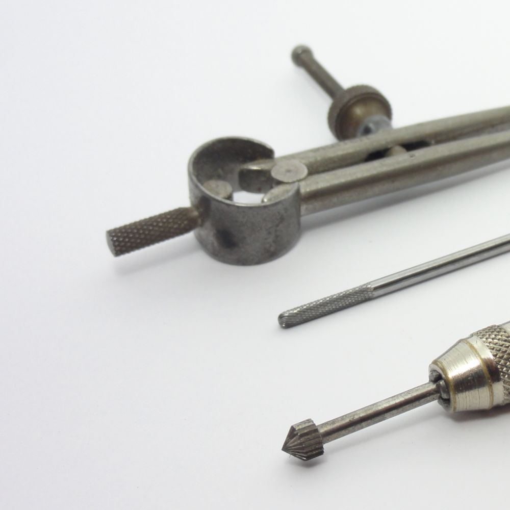 Outils de bijoutier : compas, porte-écarissoire et fraise, symbolisant la précision artisanale.